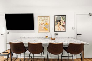 Une cuisine moderne avec un plan de travail en marbre, quatre chaises marron, un grand téléviseur à écran plat et deux œuvres d’art encadrées accrochées au mur.