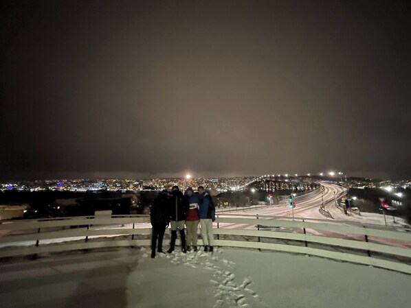 A nighttime view of a cityscape from a high vantage point, featuring a snow-covered area with a railing and a road with traffic lights. - Arctic Skyview Retreat in Tromso - With Parking (Tromsø)
