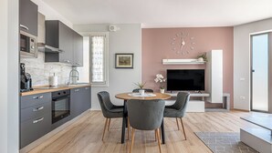 Una cocina moderna con armarios oscuros, horno empotrado y cafetera. Hay una zona de comedor con una mesa redonda y dos sillas. Un televisor de pantalla plana está montado en la pared sobre la mesa.
