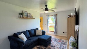 Una sala de estar con un sofá azul oscuro, un tapete estampado, un ventilador de techo y un televisor en la pared.