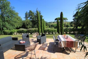 Zona de terrassa a l'exterior amb mobles de viquernia, una taula amb estovell i vista a un jardí paisatjistic amb arbres i arbustos.