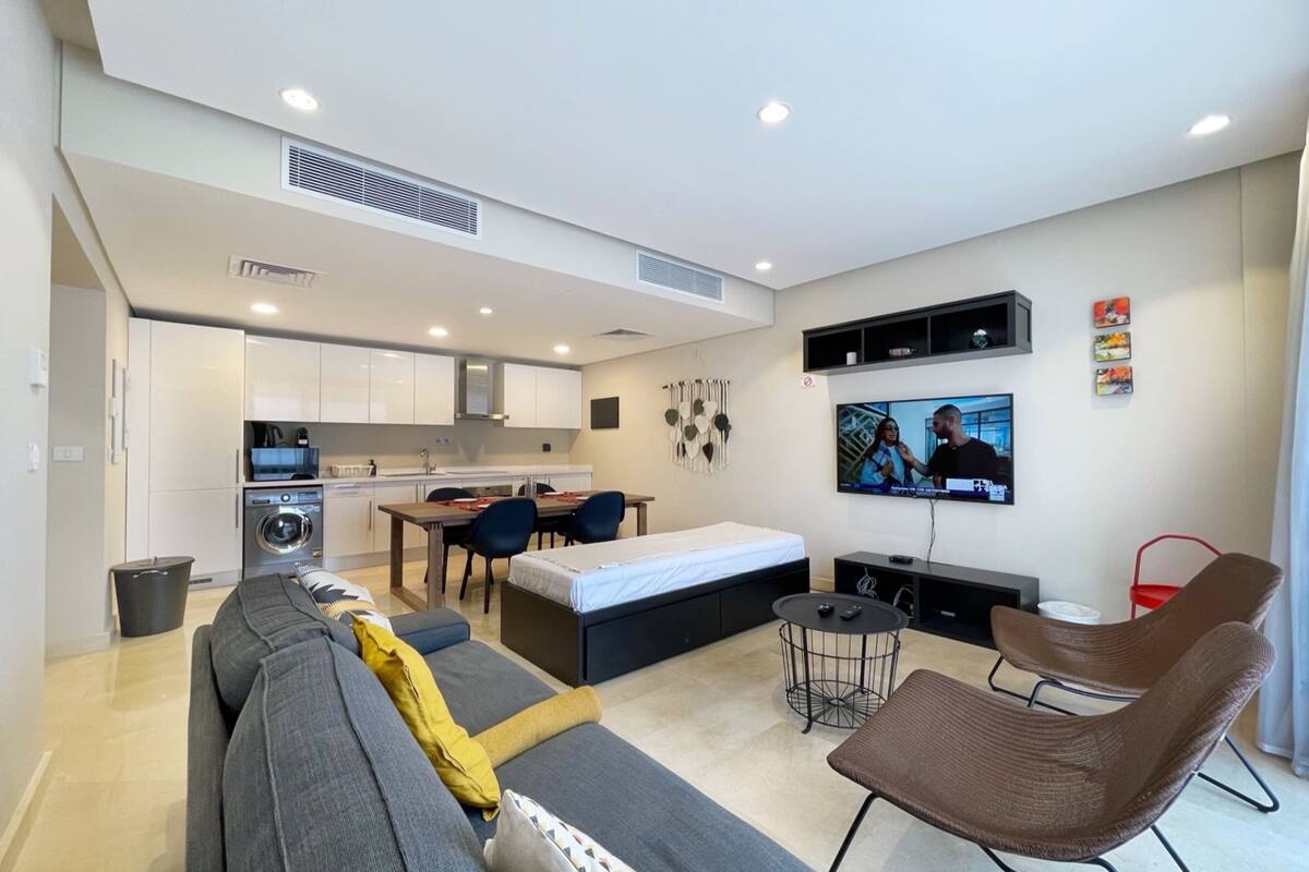 A compact living space with a sofa, a small dining table, a TV mounted on the wall, and a kitchenette in the background.