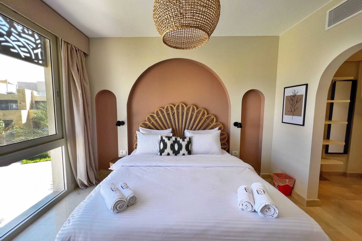 A neatly made bed with rolled towels, a large window with curtains, and a unique woven headboard.