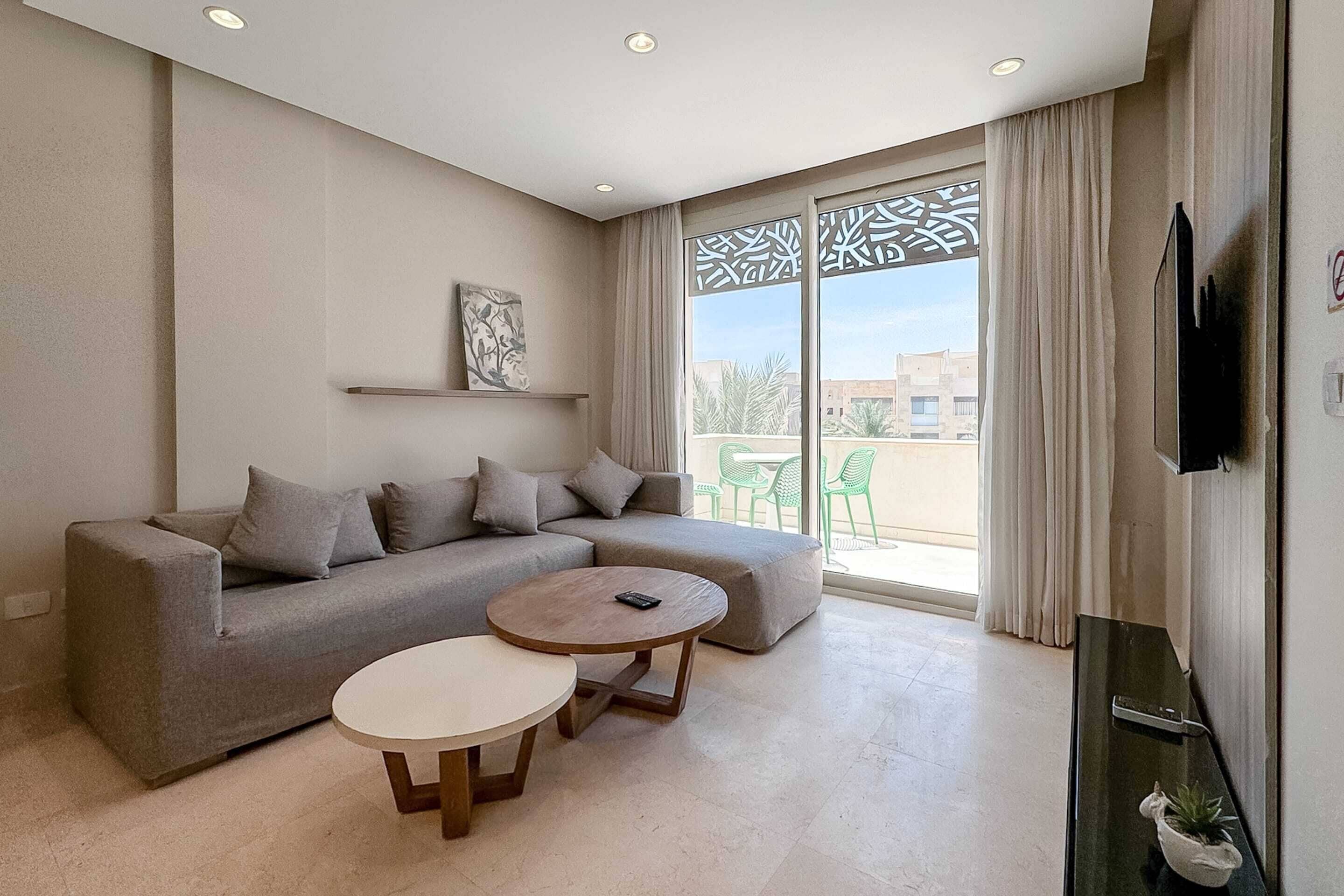 Una sala de estar moderna con un sofá seccional gris, dos mesas redondas de madera, un televisor de pantalla plana y un balcón con vistas a edificios y follaje.