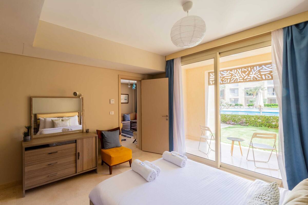 A hotel room with a bed, a wooden dresser, a small table, a chair, and a view of an outdoor area with a pool and greenery.