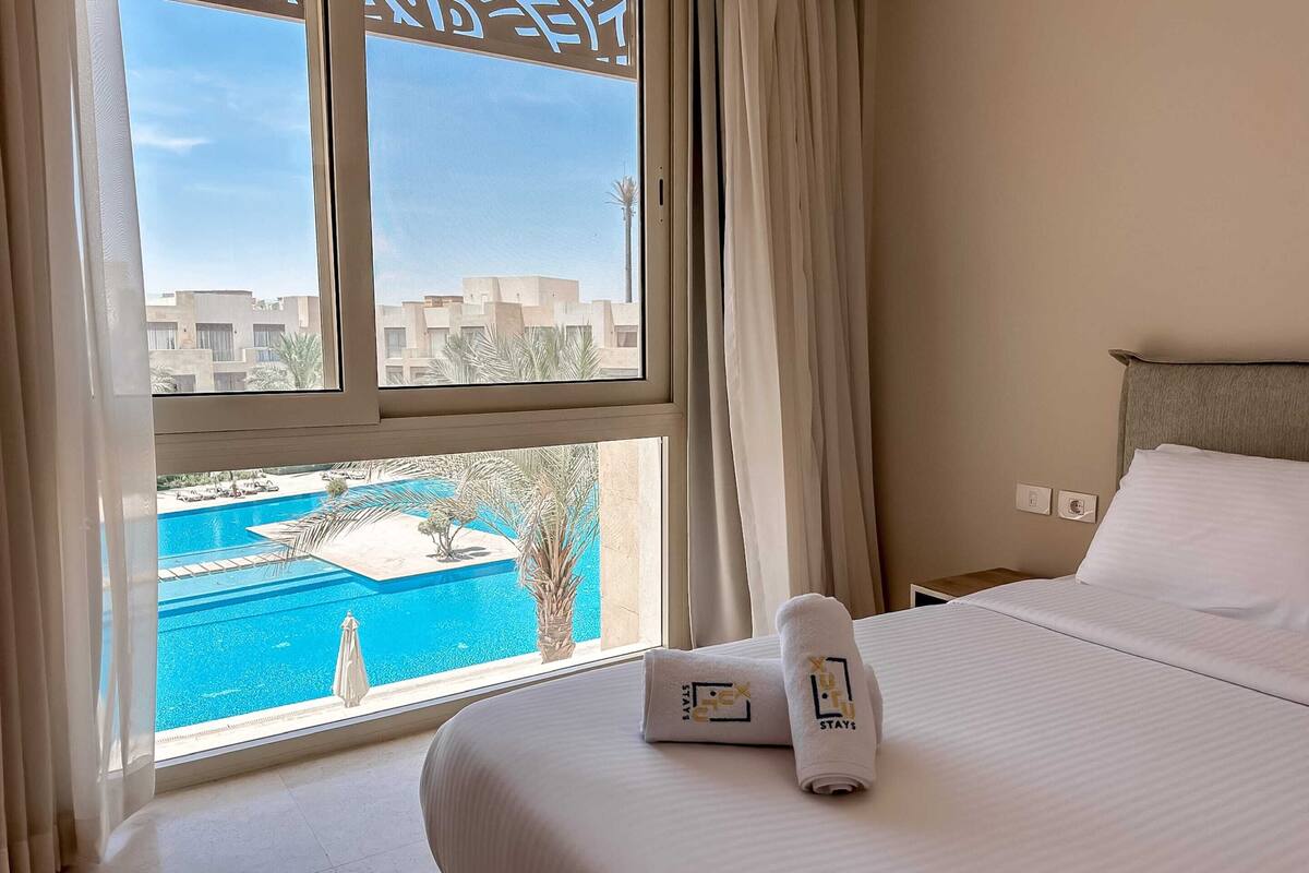 A hotel room with a bed, a window with curtains, a view of a swimming pool, and a towel with a logo.