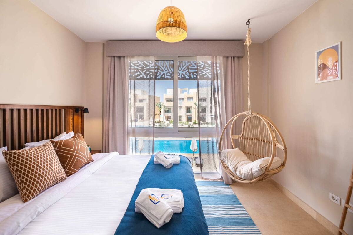 A hotel room with a bed, a hanging chair, a view of a pool, and a large window with patterned curtains.