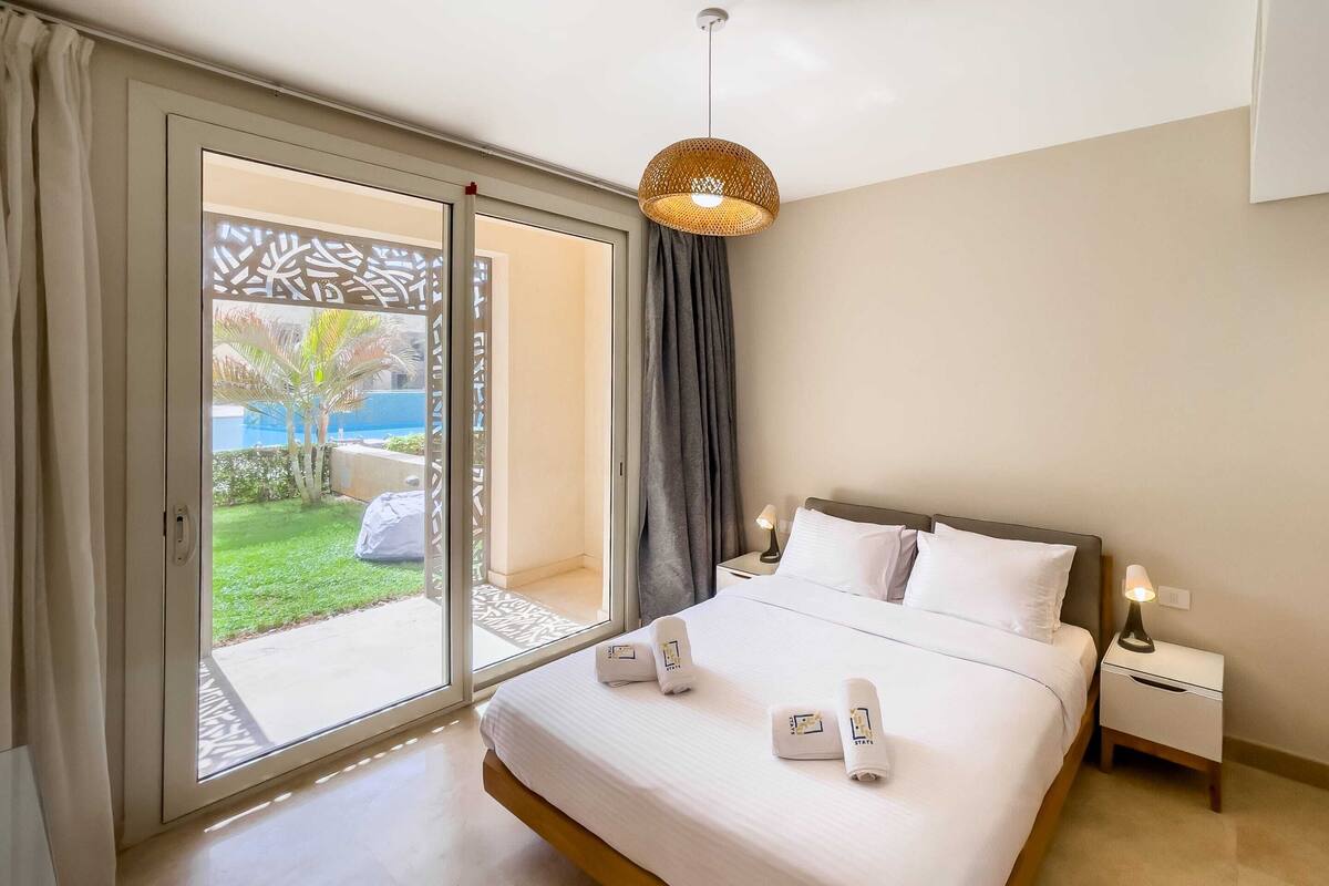 A hotel room with a bed, a nightstand, a lamp, and a view of a garden through a sliding door.