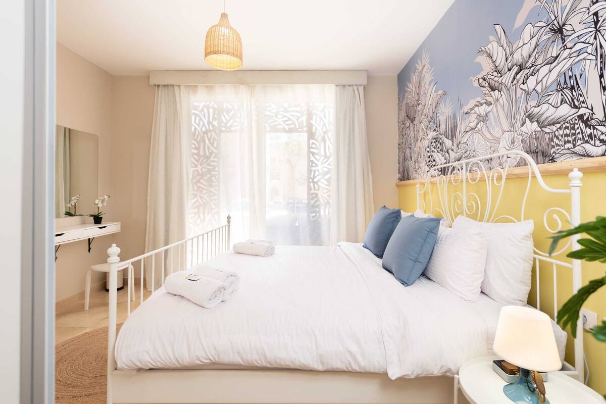A bedroom with a bed, a bedside table with a lamp, a small table with a plant, and a wall with a botanical mural.