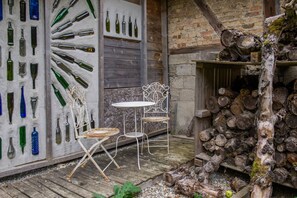 Un espace extérieur aménagé dans un style rustique, avec des chaises et une table en métal, entouré d’une terrasse en bois et d’un mur en briques.