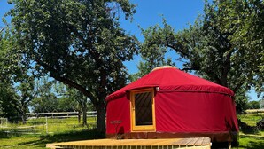 A red yurt with a wooden door, a wooden platform, and a fenced area with trees.