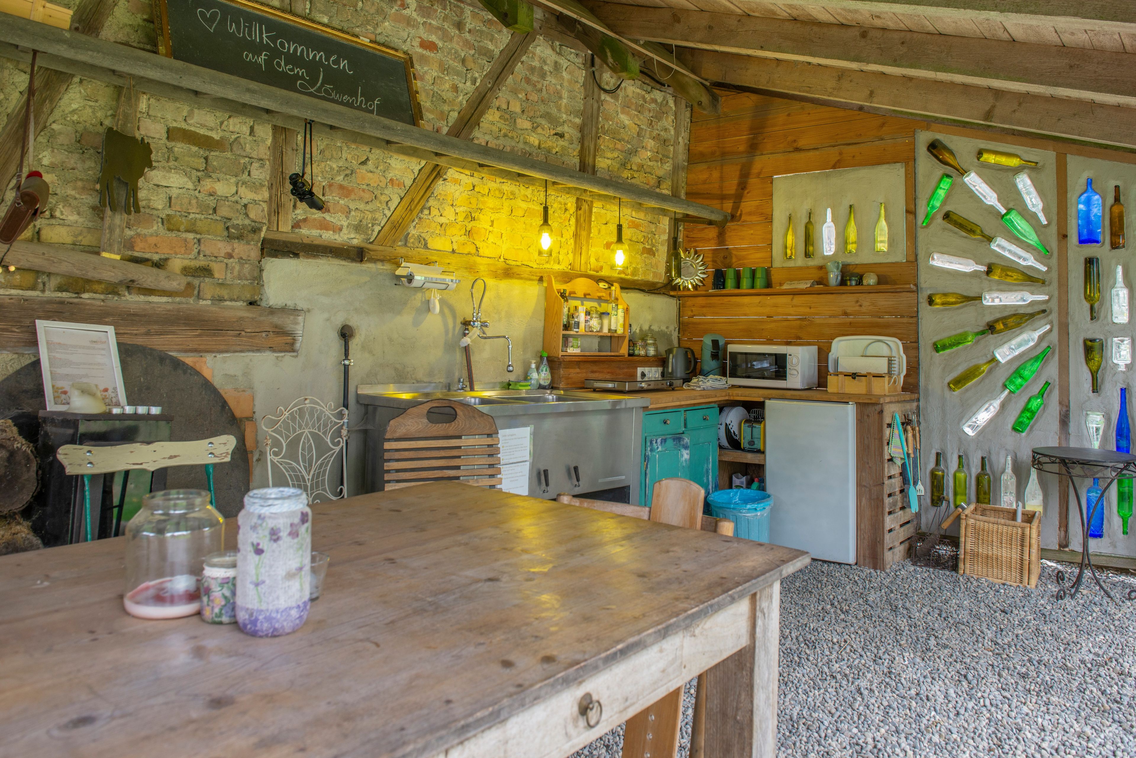 Een landelijke keuken met een houten tafel, een schoolbord met een welkomstboodschap, en een muur versierd met lege flessen.