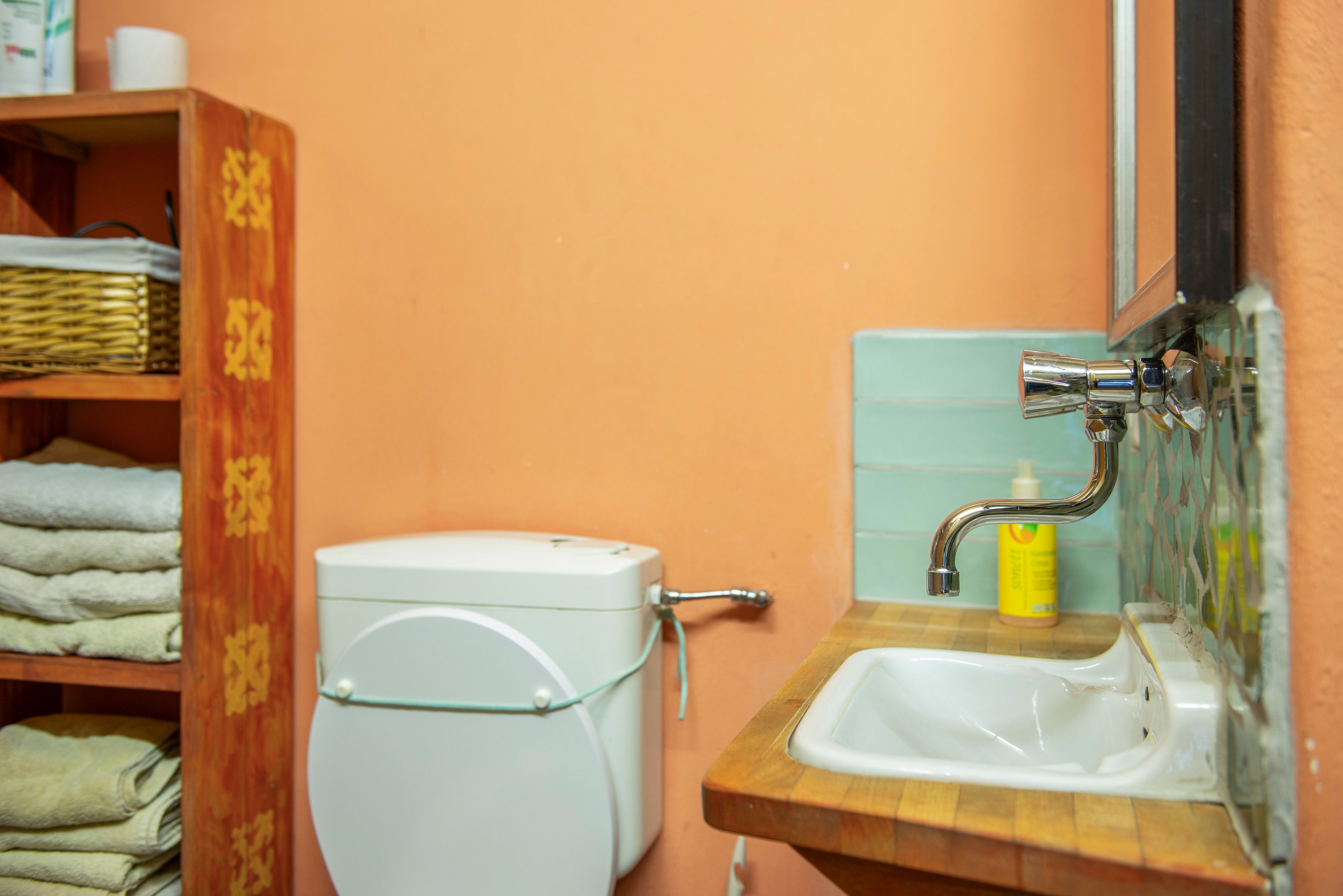 Un baño con inodoro blanco, un estante de madera con toallas dobladas, un lavamanos con grifo y una botella de jabón.