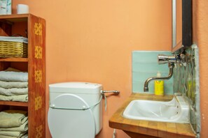 Une salle de bain avec des toilettes blanches, une étagère en bois où sont pliées des serviettes, un lavabo avec un robinet et un flacon de savon.