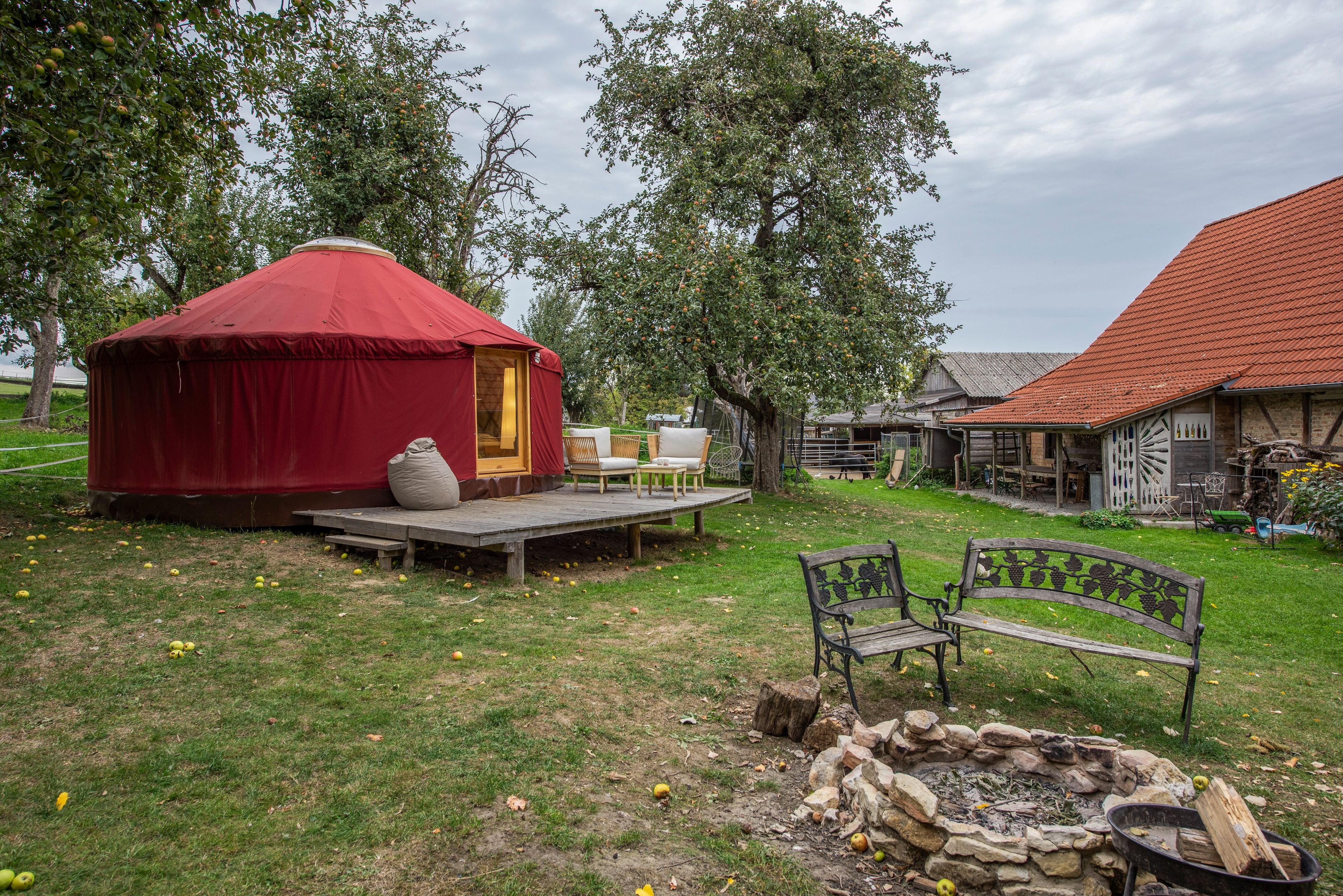 A yurt with a wooden deck, a stone fire pit, and a rustic building with a red roof.