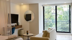 A modern hotel room with a bed, a sofa, a small table, a mirror, a TV, and a large window offering a view of greenery. - NOK Cozy Studio in Parque Virrey (Bogotá)