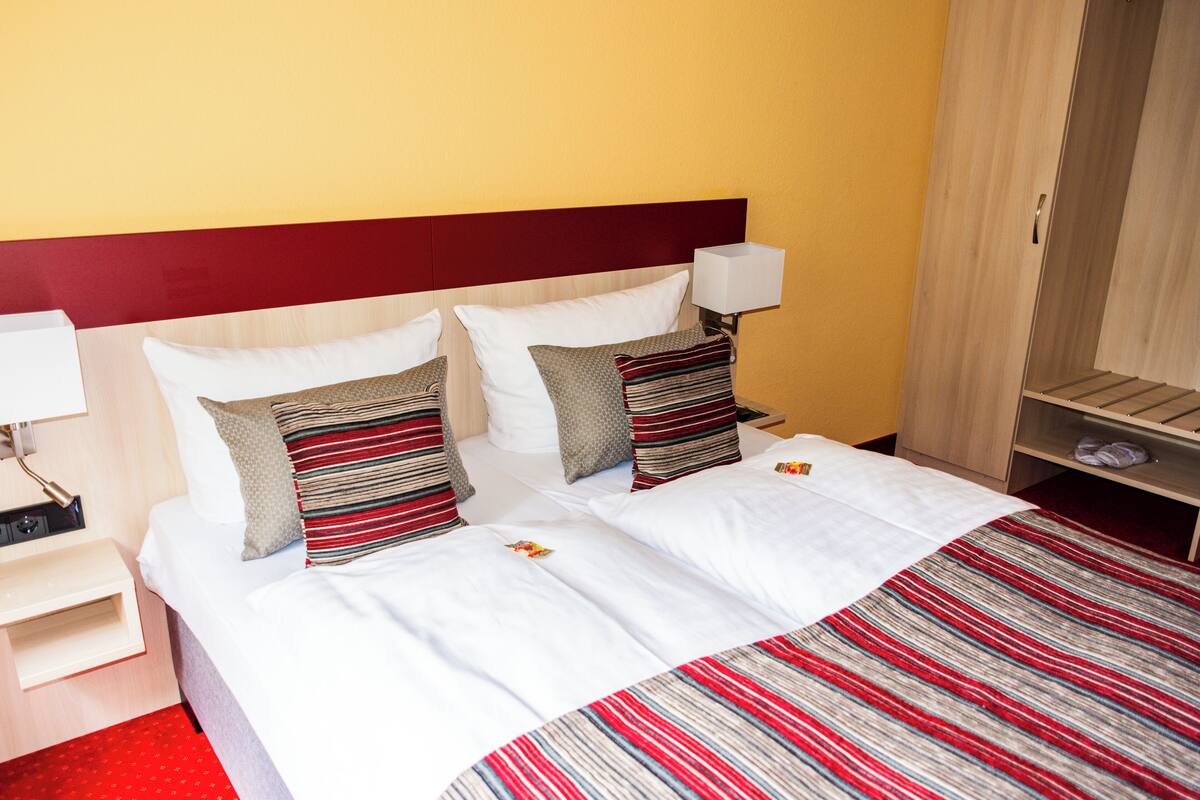 Una habitación de hotel con una cama, almohadas, una mesita de noche, una lámpara y un armario.