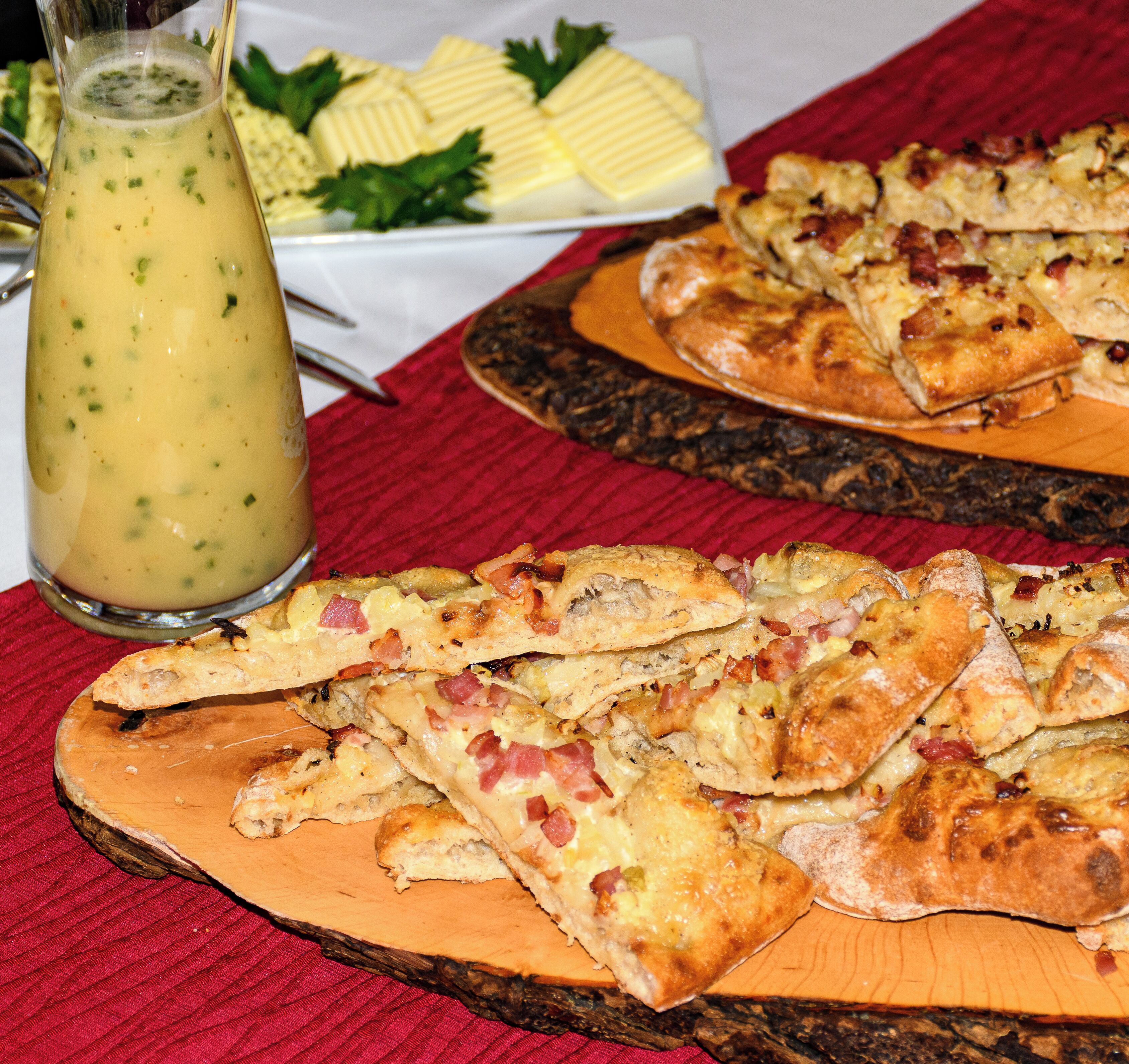 Une table sur laquelle se trouvent une planche en bois servant de présentoir pour des parts de pizza, un pichet de soupe et une assiette avec du beurre et des légumes verts.
