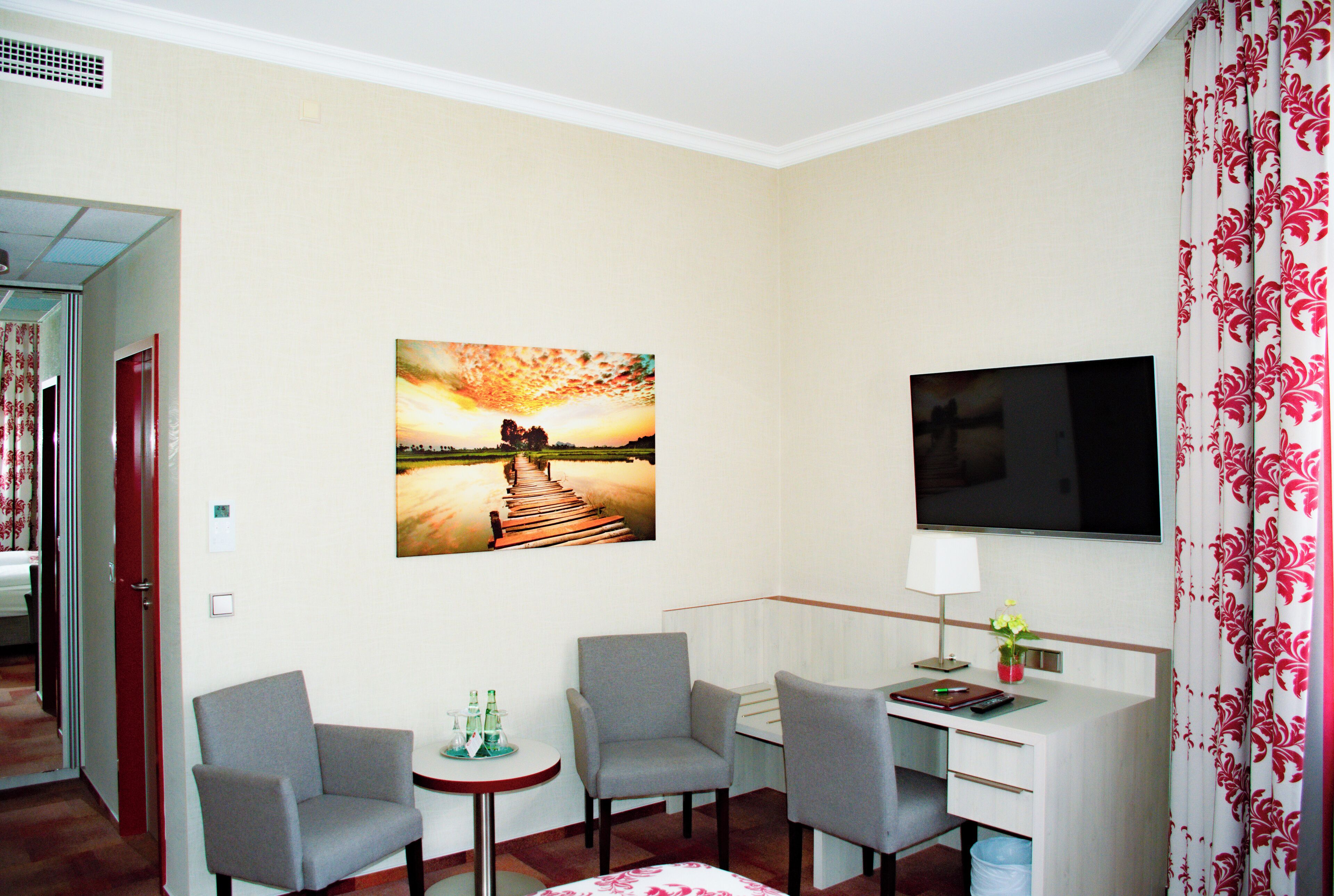 A hotel room with a TV, two chairs, a small table with drinks, and a painting of a sunset over a pier.