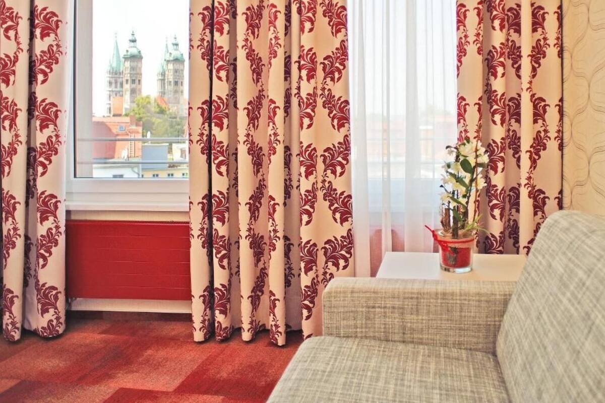 Una habitación con ventana, radiador rojo, cortinas beige con estampado floral, un sofá beige y una mesita con planta en maceta.