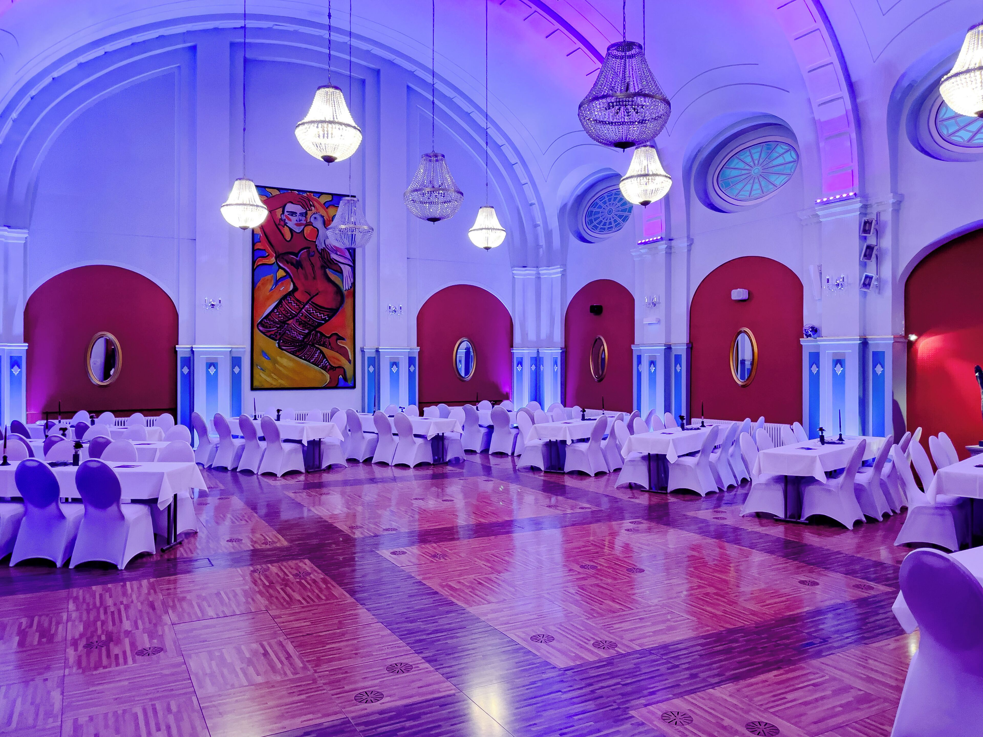 Ein Festsaal mit runden Tischen, Kronleuchtern und einem großen Gemälde an der Wand.