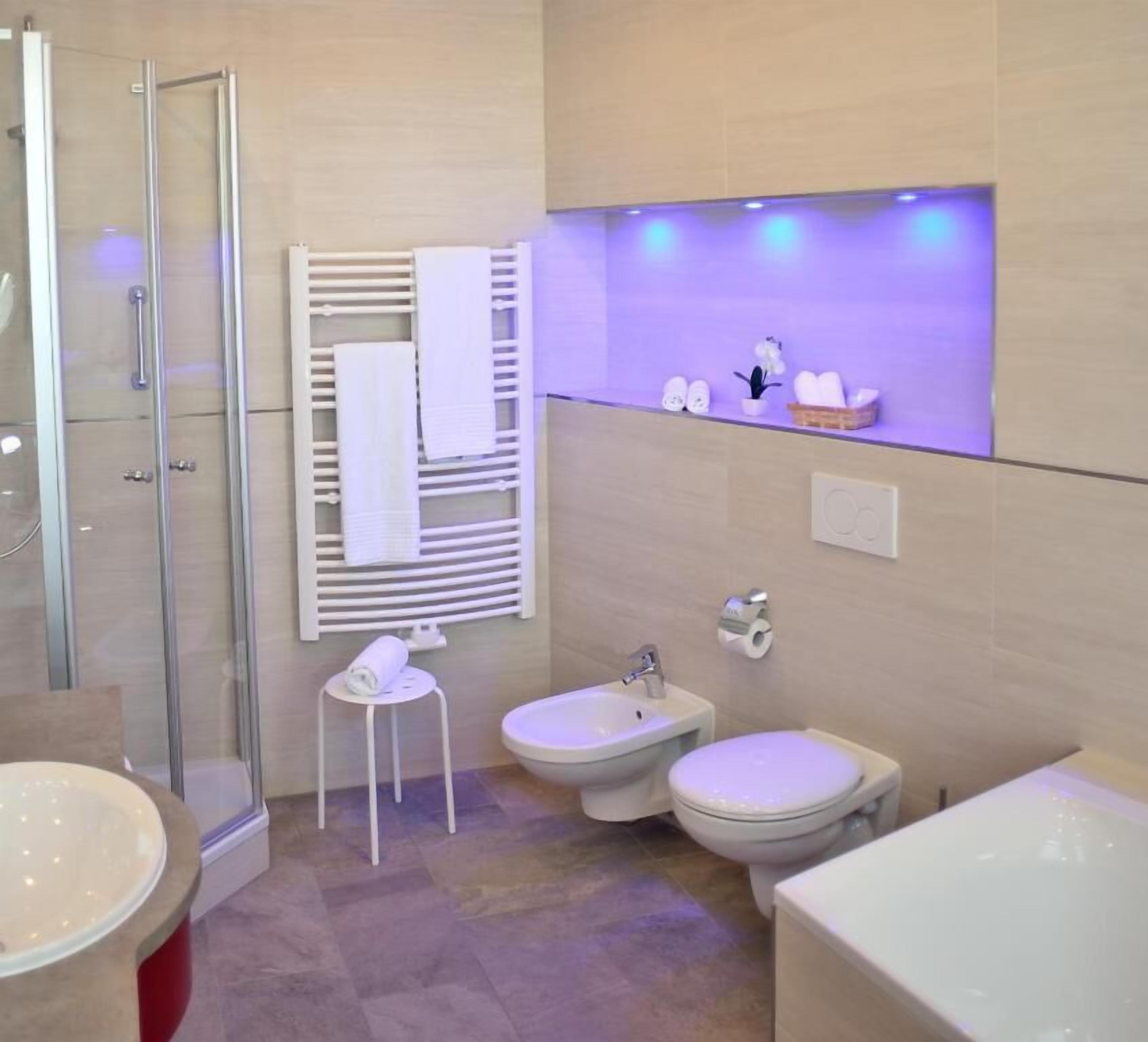 A modern bathroom with a glass shower enclosure, a small round table with a towel, two bidets, and a vanity with a sink and mirror.
