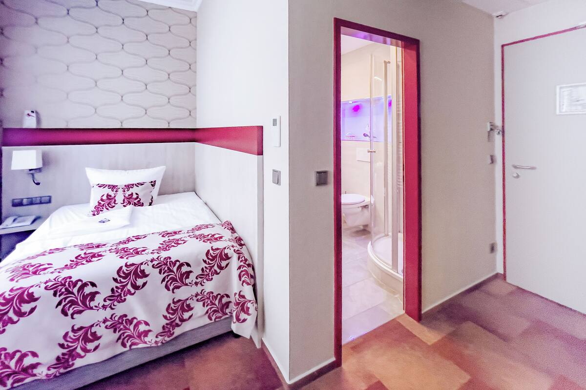 Habitación de hotel con cama, mesita de noche, baño a la vista a través de una puerta abierta y puerta con marco rojo.