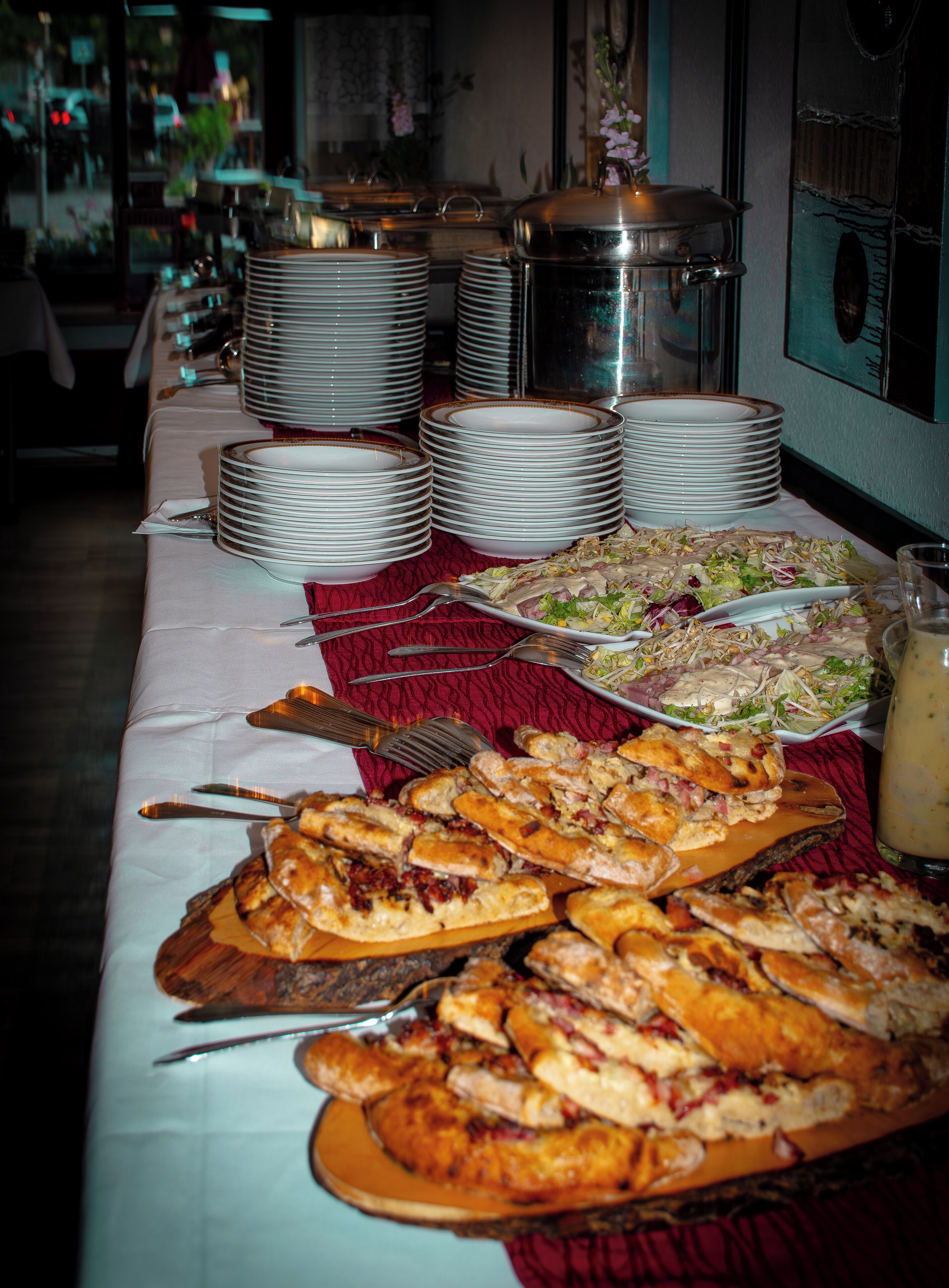 A buffet table with various dishes, including pizzas and salads, and stacks of plates and silverware.
