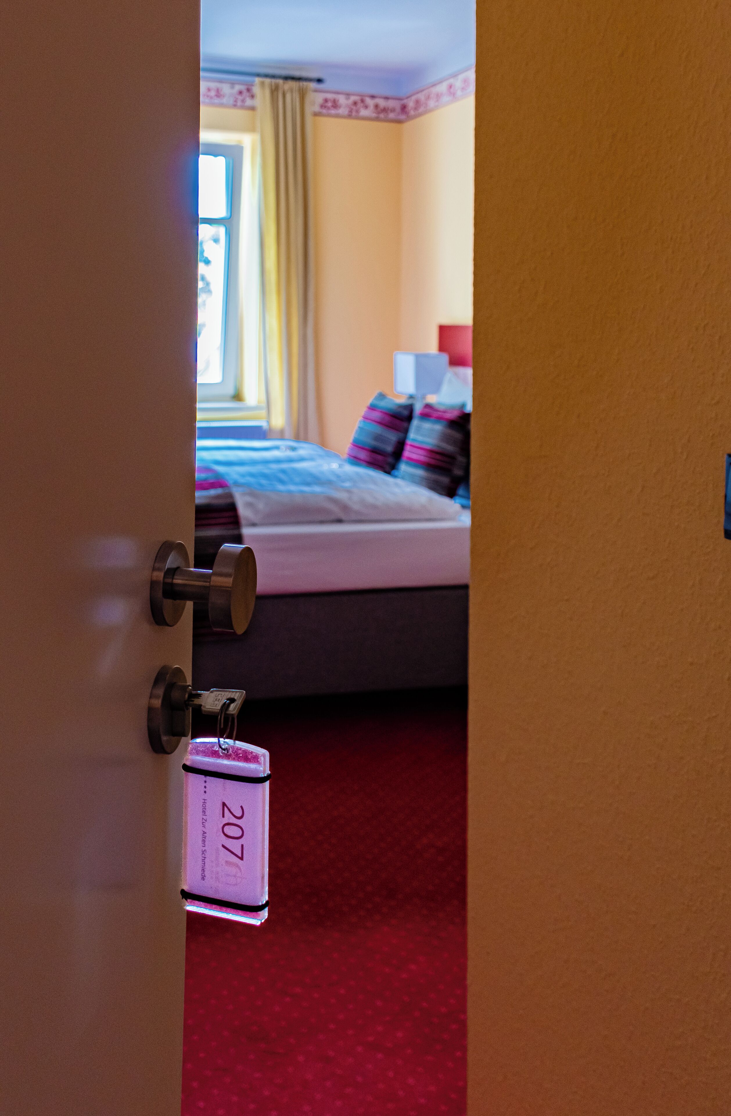 A hotel room with a bed, a red carpet, and a door with a keycard lock.