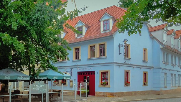 Un bâtiment bleu avec une porte rouge, portant l’inscription « RESTAURANT », et une enseigne indiquant « BUDBRAMBACHER ».