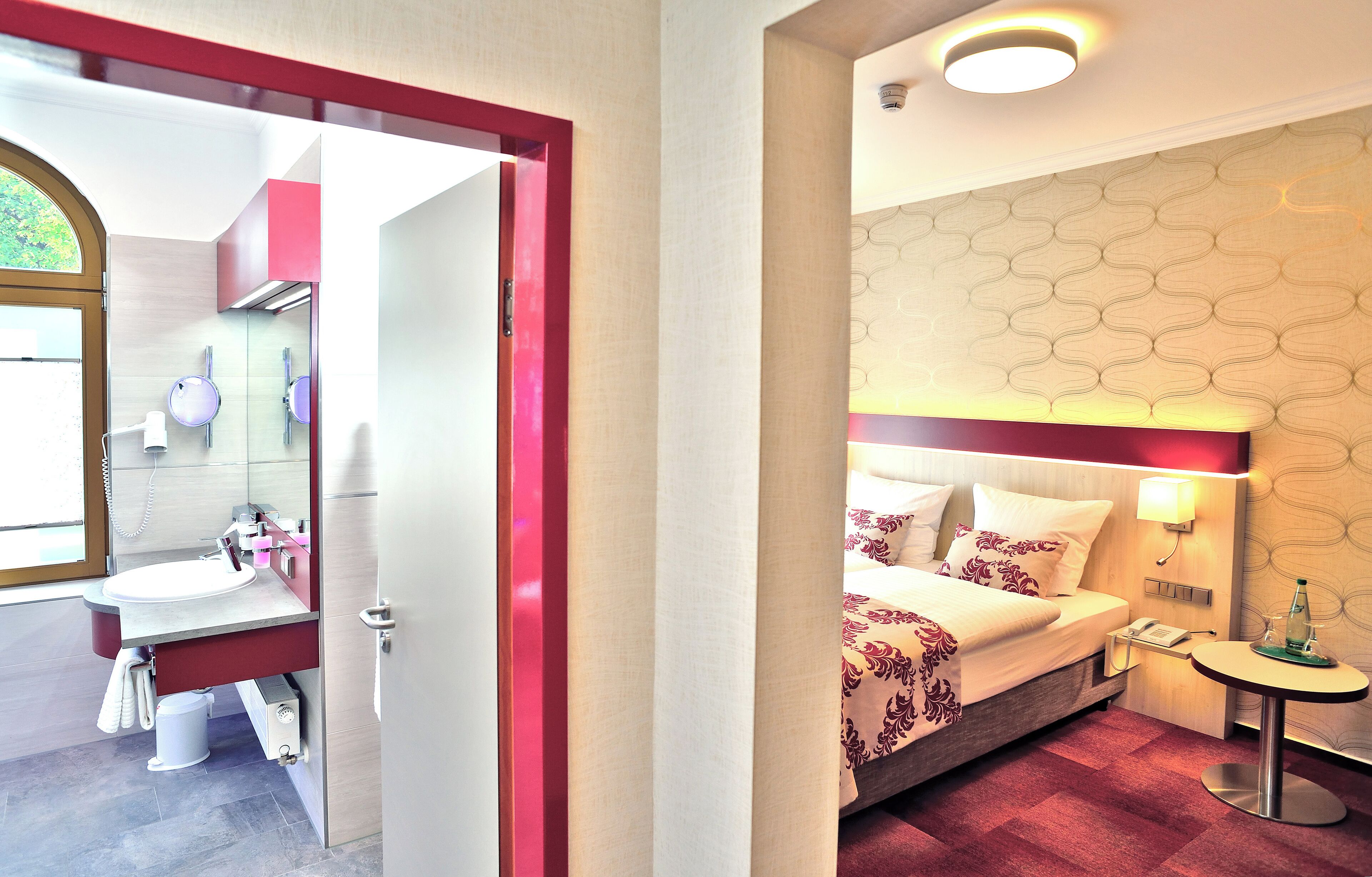Una habitación de hotel con cama, mesita de noche y baño con lavamanos y espejo.