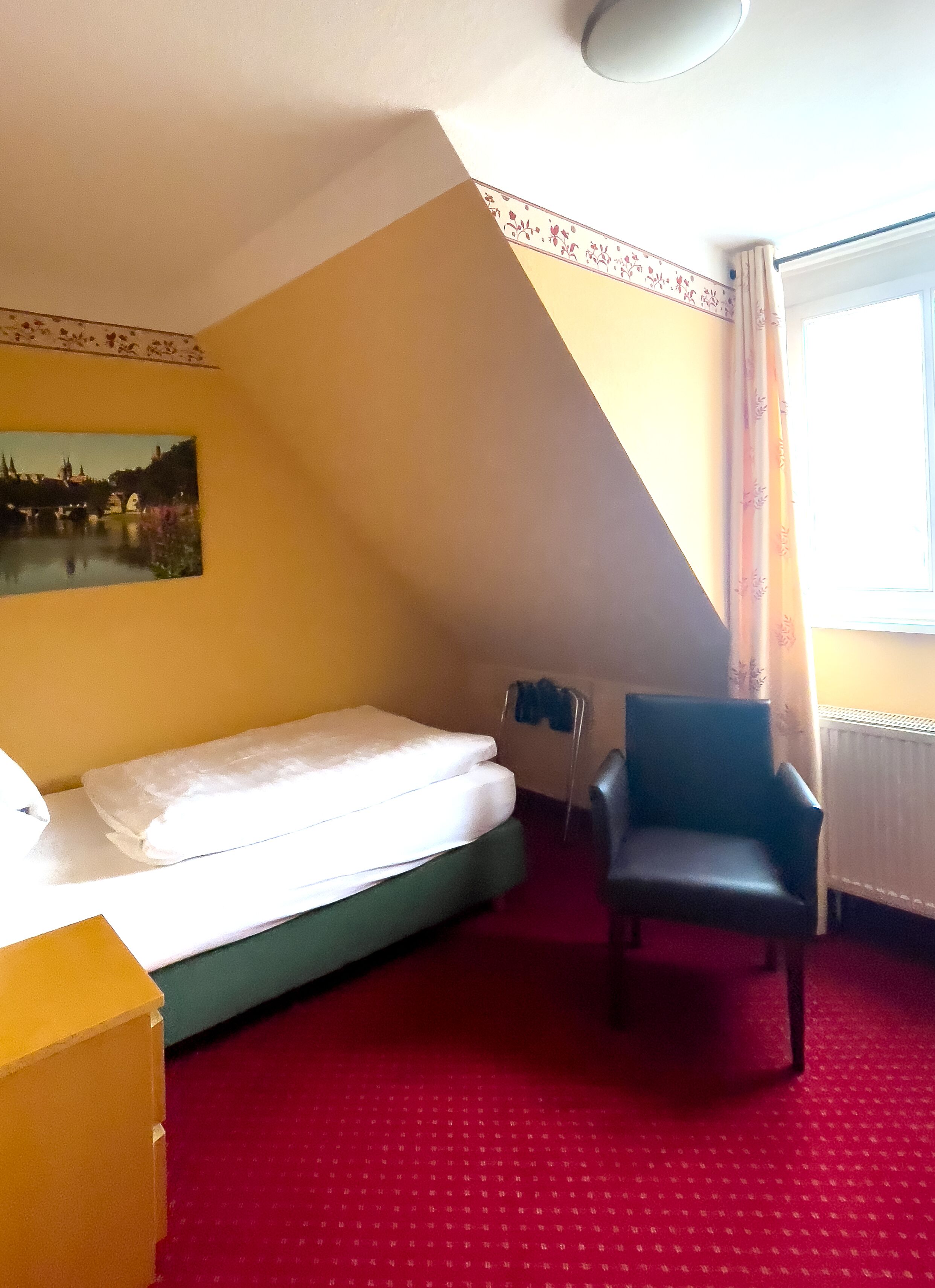 Een hotelkamer met een bed, een stoel, een schilderij aan de muur en een raam met gordijnen.