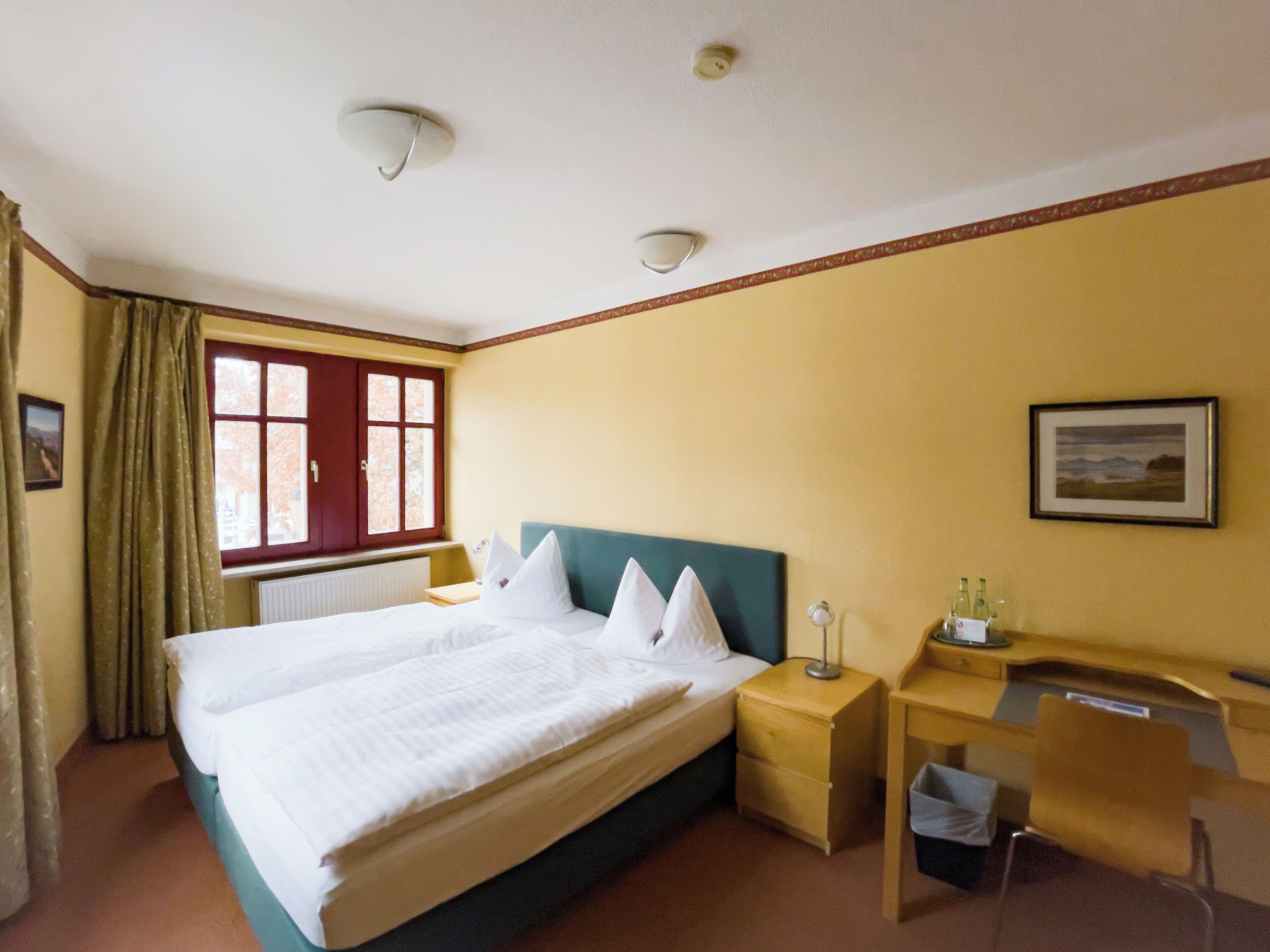 Une chambre d’hôtel comprenant un lit, une table de chevet, un bureau, une chaise, une fenêtre avec des rideaux et un tableau au mur.