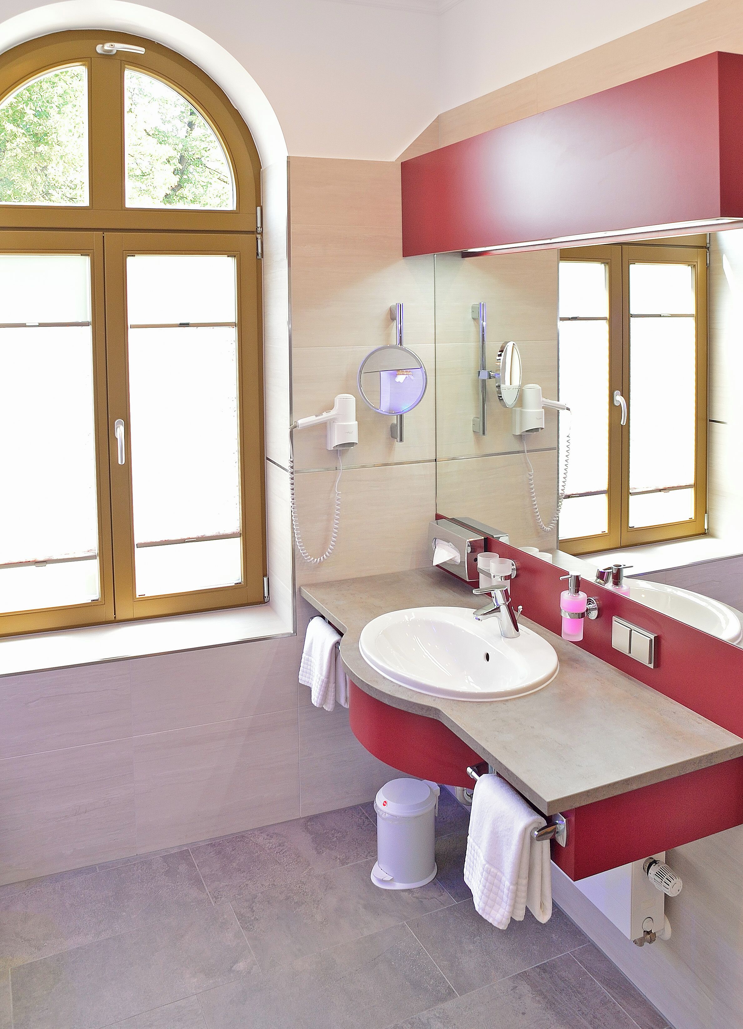 A bathroom with a large window, a round mirror, a sink, and a red vanity.