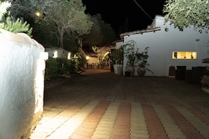Un patio empedrado con un edificio blanco, plantas en macetas y mesas al aire libre.