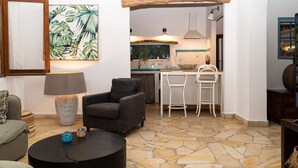 Un salon avec un sol en pierre, une table basse noire, un canapé gris et un tableau représentant des feuilles tropicales.