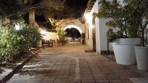 Une cour dallée avec un chemin en pierre, des plantes en pot et un bâtiment blanc doté d’une entrée en arc de cercle.