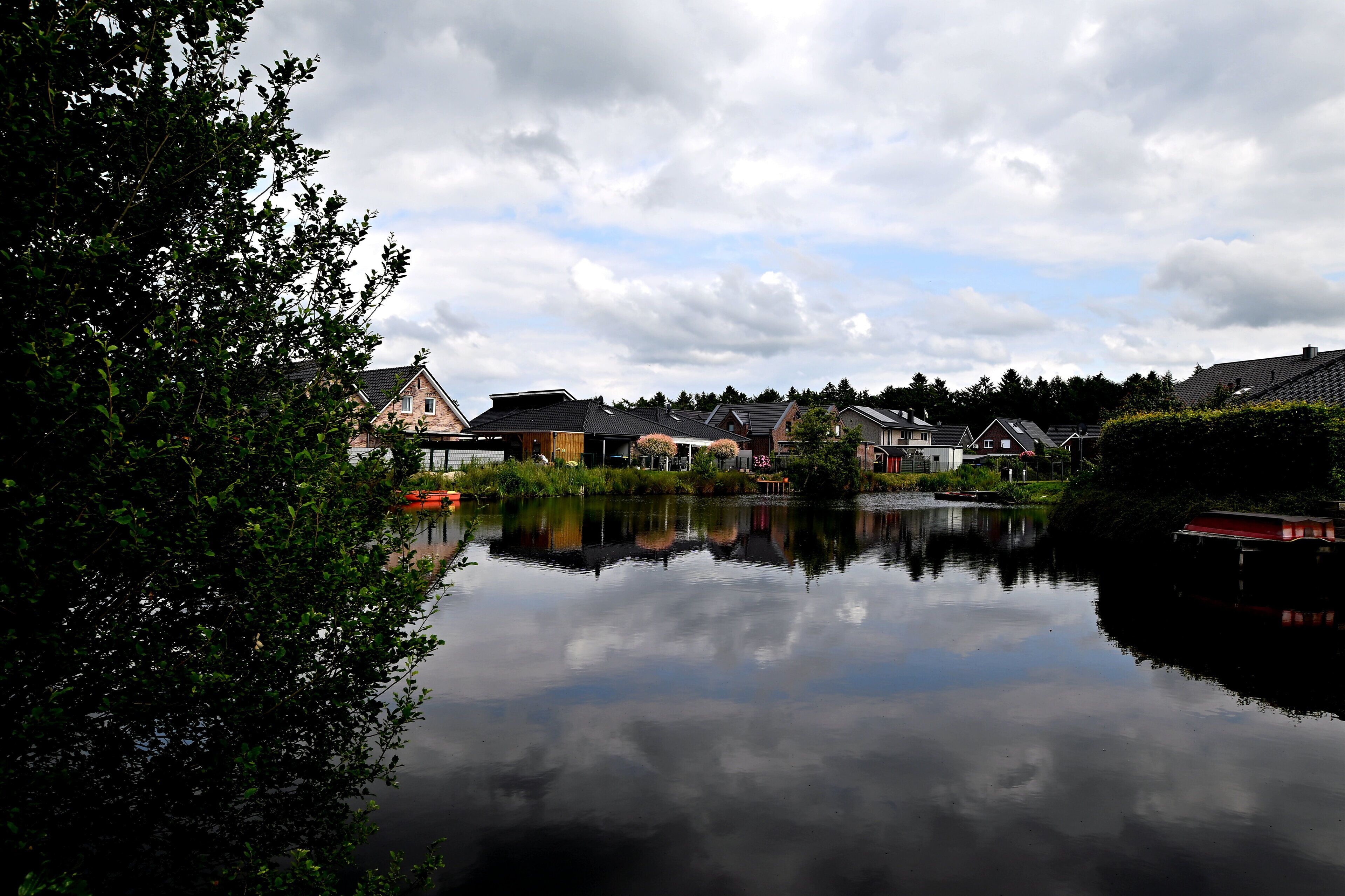 A calm lake reflecting houses and trees, surrounded by greenery.