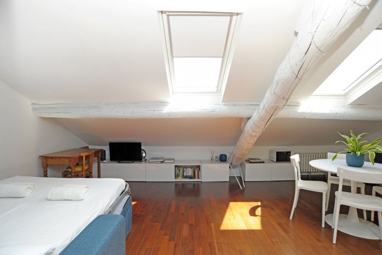 A compact room with a loft bed, a desk, a chair, a small table, and a potted plant.