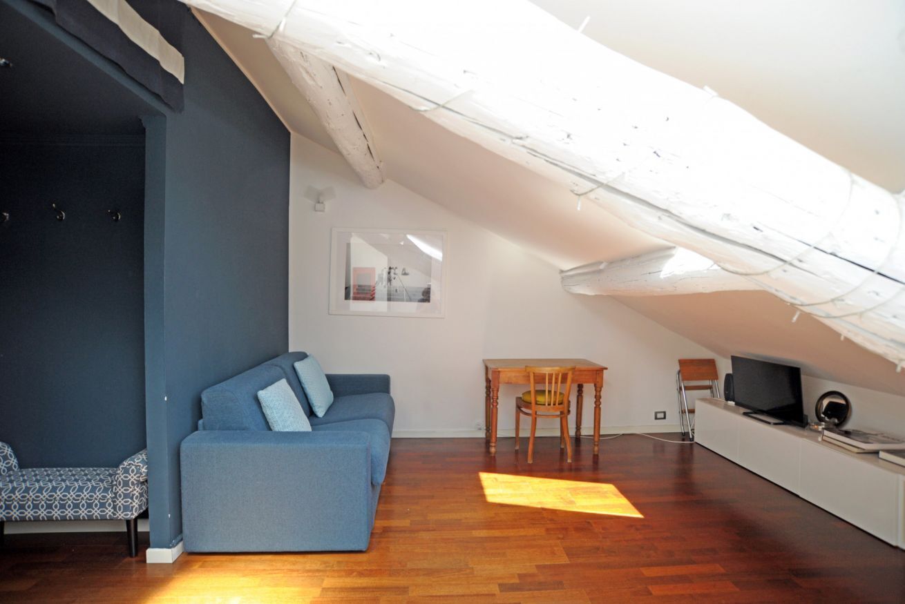 Une chambre mansardée avec un canapé bleu, une table à manger en bois, un téléviseur et un plafond blanc.