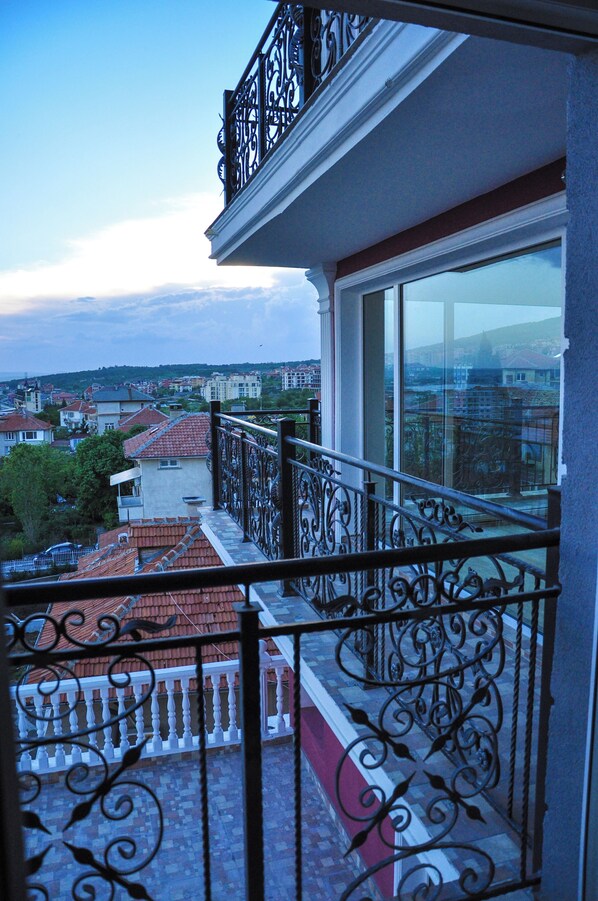 A balcony with wrought iron railings overlooking a cityscape with red-roofed buildings and a clear sky.