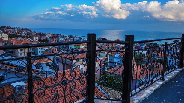 A coastal cityscape with red-tiled rooftops, a sea view, and a balcony with ornate black iron railings.