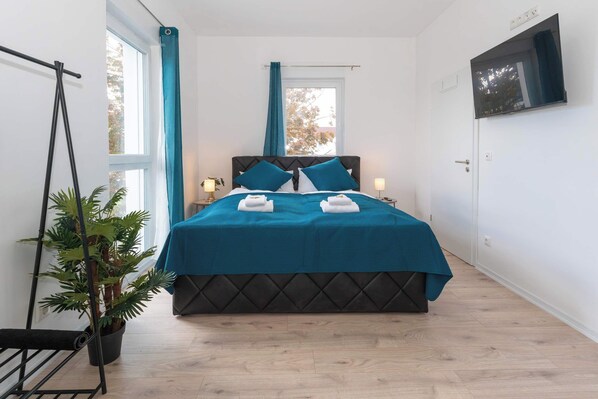 Une chambre à coucher avec un lit, une plante en pot, une fenêtre avec des rideaux et un téléviseur fixé au mur.