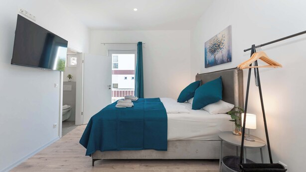 A modern bedroom with a bed, a TV, a bedside table with a lamp, a hanging rack, and a view of the outside. - LiT LiVING: Luxus | Box SprIng | Kitchen | Parking (Altenstadt)