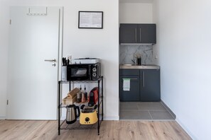 A compact kitchen with a microwave, toaster, and kettle on a shelf, a small refrigerator, and a dark cabinet.