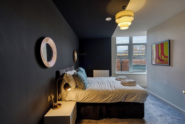 A modern bedroom with a bed, bedside table, lamp, and artwork. - Renaissance House Apartments (Southport)