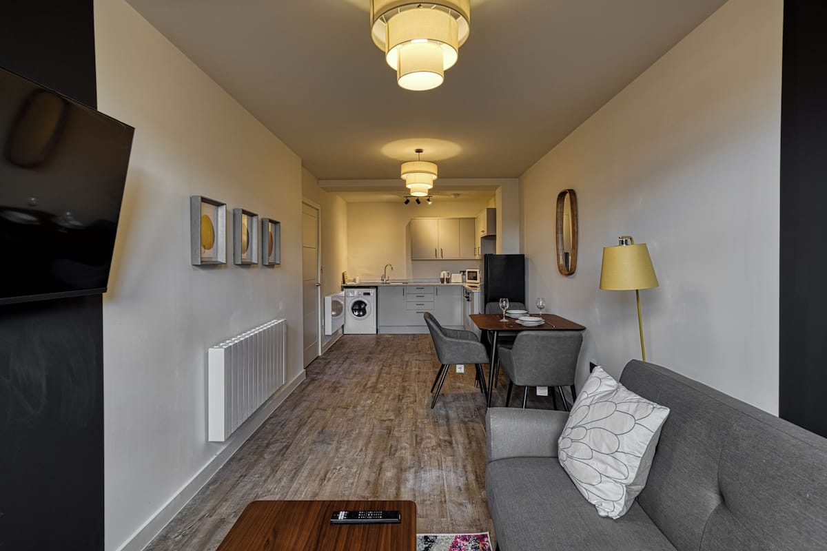 Un appartement moderne comprenant un salon, un coin repas et une cuisine. Il est équipé d’un canapé, d’une table à manger, de chaises, d’un téléviseur et d’une lampe.