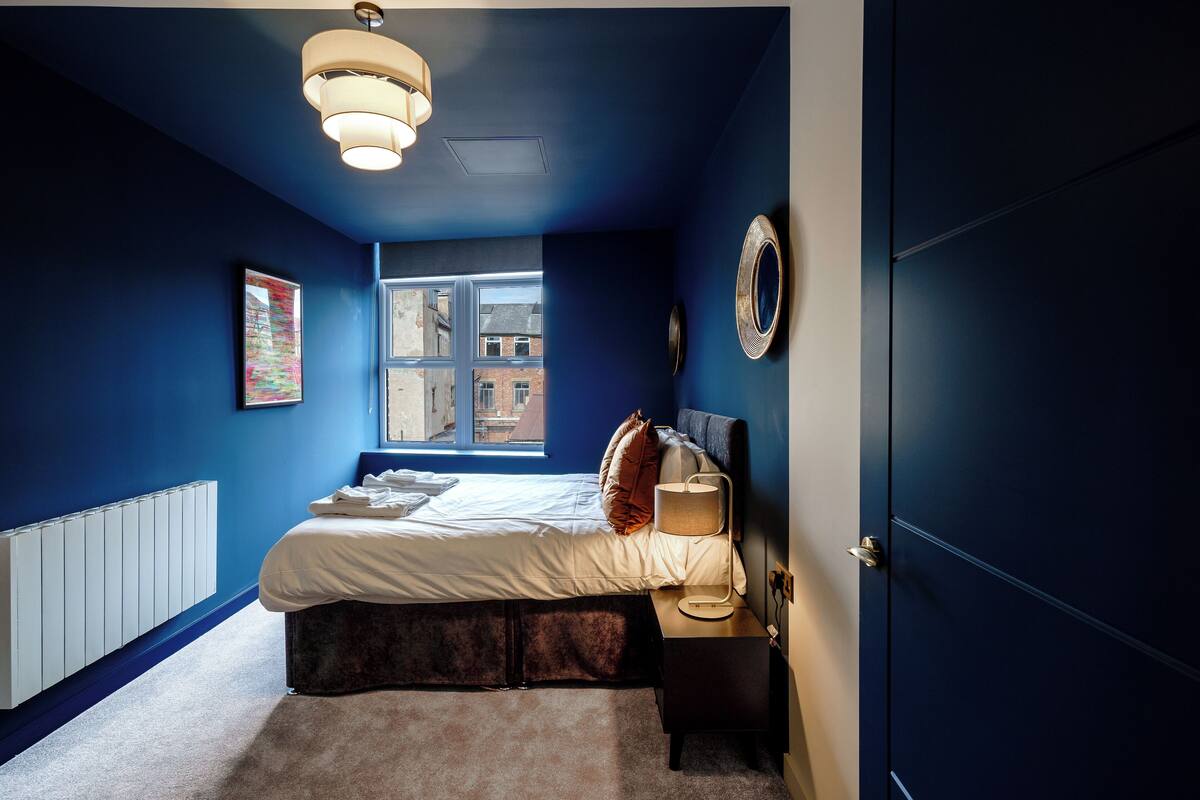 Une chambre à coucher comprenant un lit, une table de chevet, une lampe, une fenêtre, un radiateur et une œuvre d’art accrochée au mur.