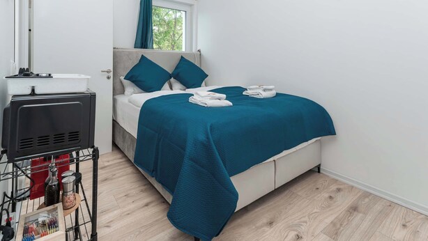 A hotel room with a bed, a window with curtains, a bedside table with a lamp, and a wall-mounted heater. - LiT LiVING: Luxus | Box SprIng | Kitchen | Parking (Altenstadt)