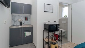 A compact kitchen with grey cabinets, a microwave, a toaster, and a small shelf with kitchen items. - LiT LiVING: Luxus | Box SprIng | Kitchen | Parking (Altenstadt)